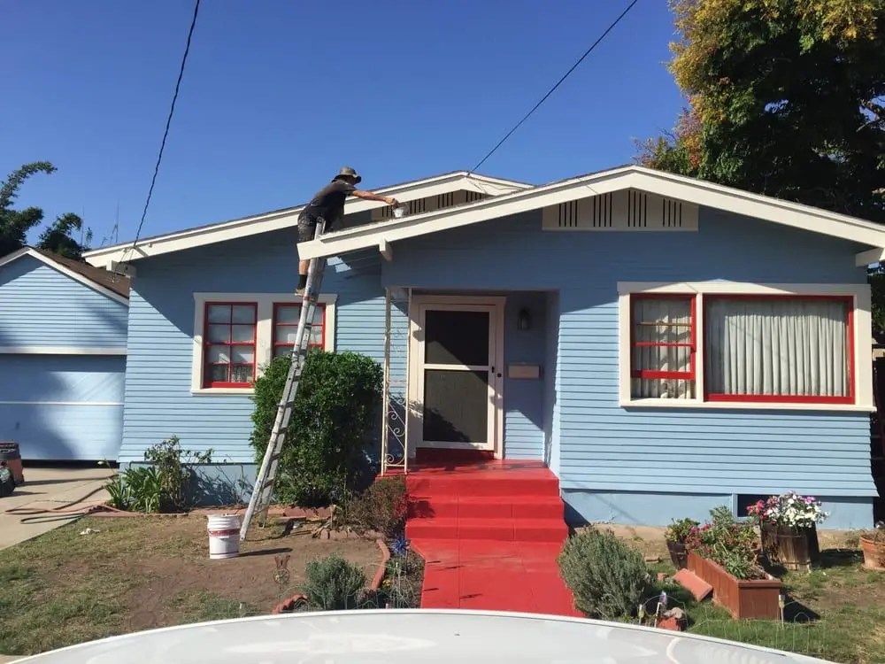 A Montgomery painter on a ladder painting the eaves of a light blue Chula Vista bungalow with red porch steps